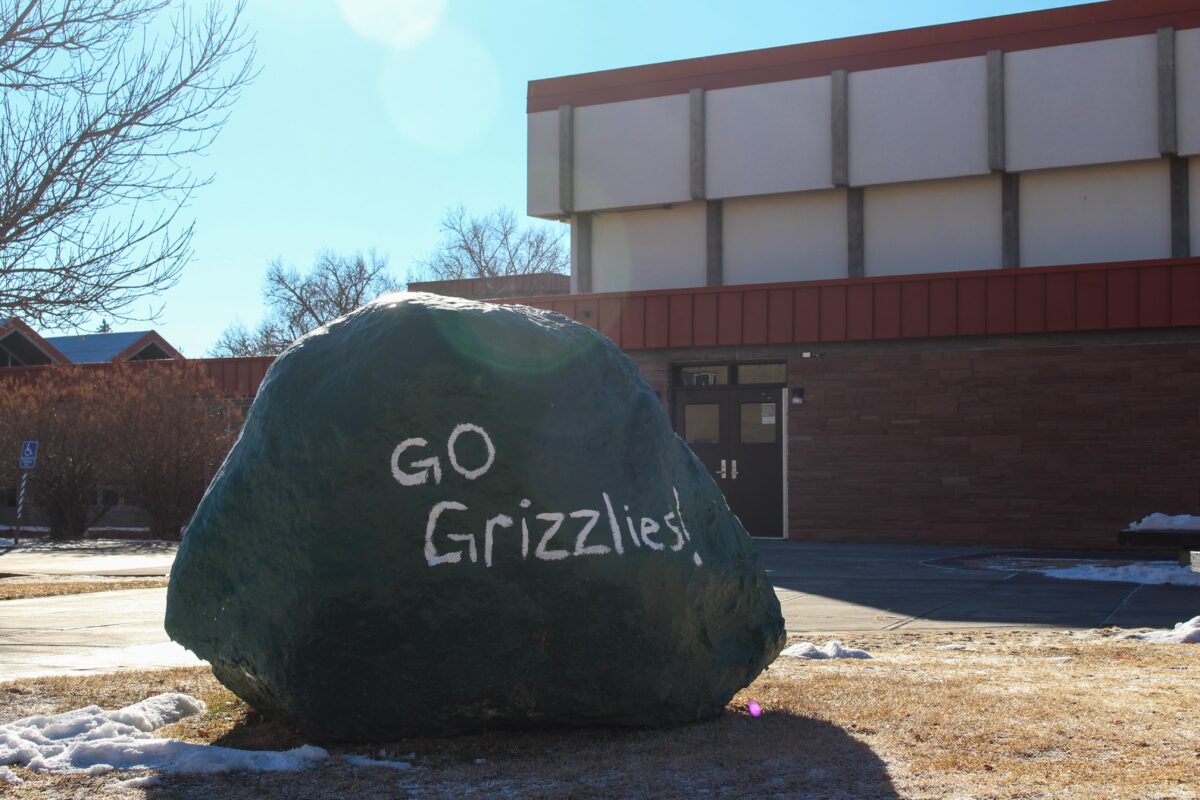 A boulder is painted green with "Go Grizzlies!" written in white near the Student Union Building on the Adams State campus.